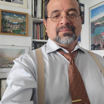 Antonio Tovar wears round, metal rimmed glasses, a button-down shirt, and a brown tie and suspenders. He is standing in front of a wall that has colorful paintings on it next to a built-in bookcase.