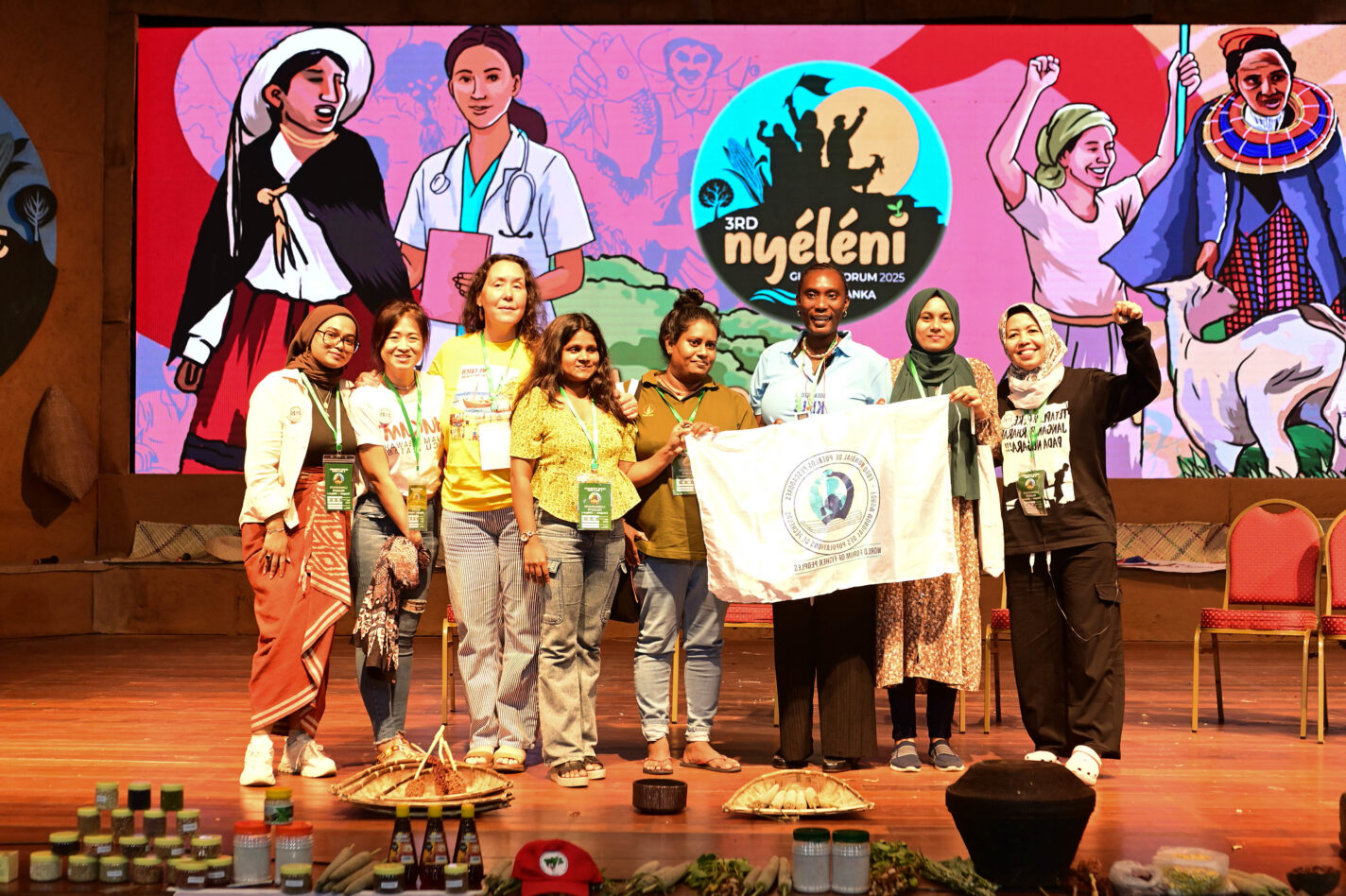 Eight diverse members of the Women's Assembly at the 3rd Nyéléni Global Forum in Kandy, Sri, Lanka, stand on stage together. There is a colorful digital image of diverse women from all over the world in the background.