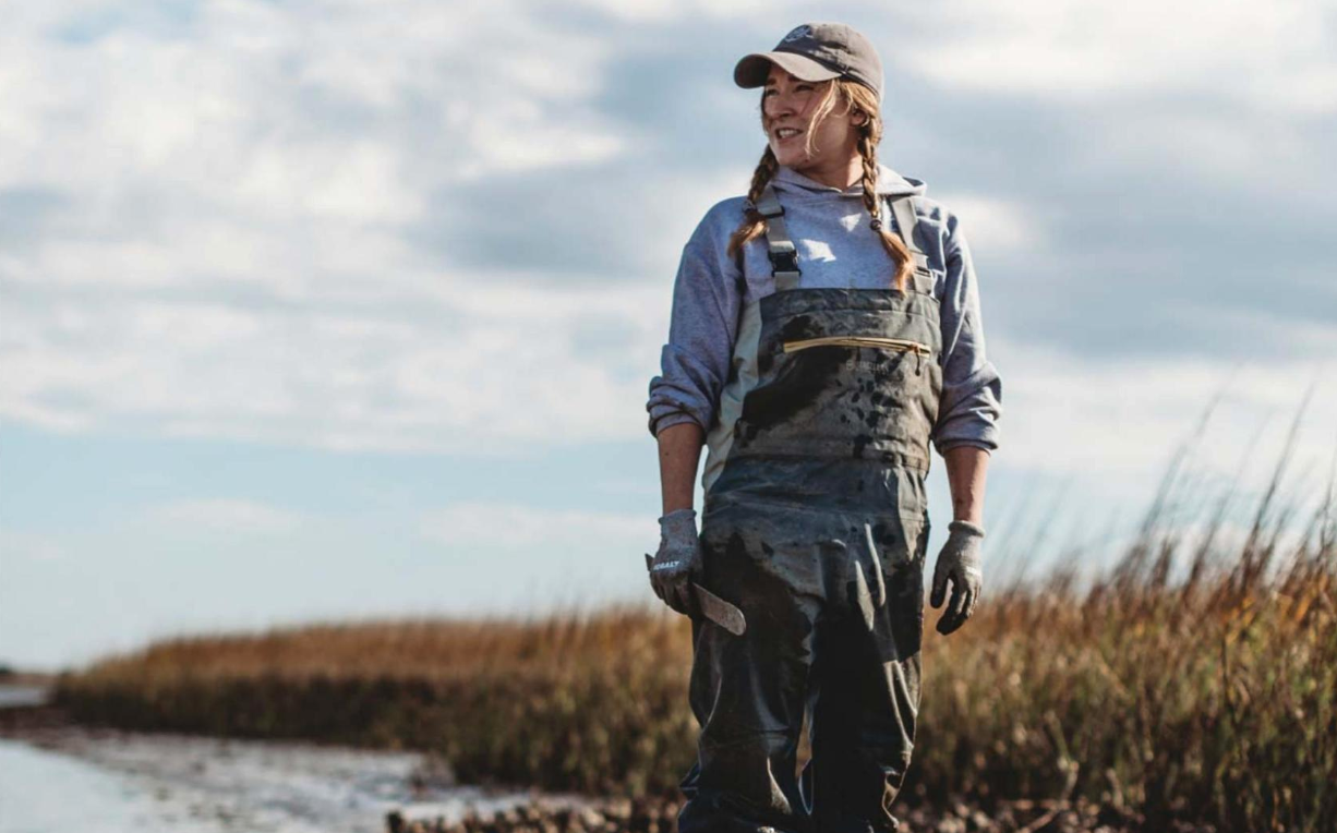Fisherwoman Ana Shellem stands in a marshy area with a partly cloudy sky behind her. She is wearing a baseball cap and suspenders and is smiling. A muddy bucket sits in front of her.