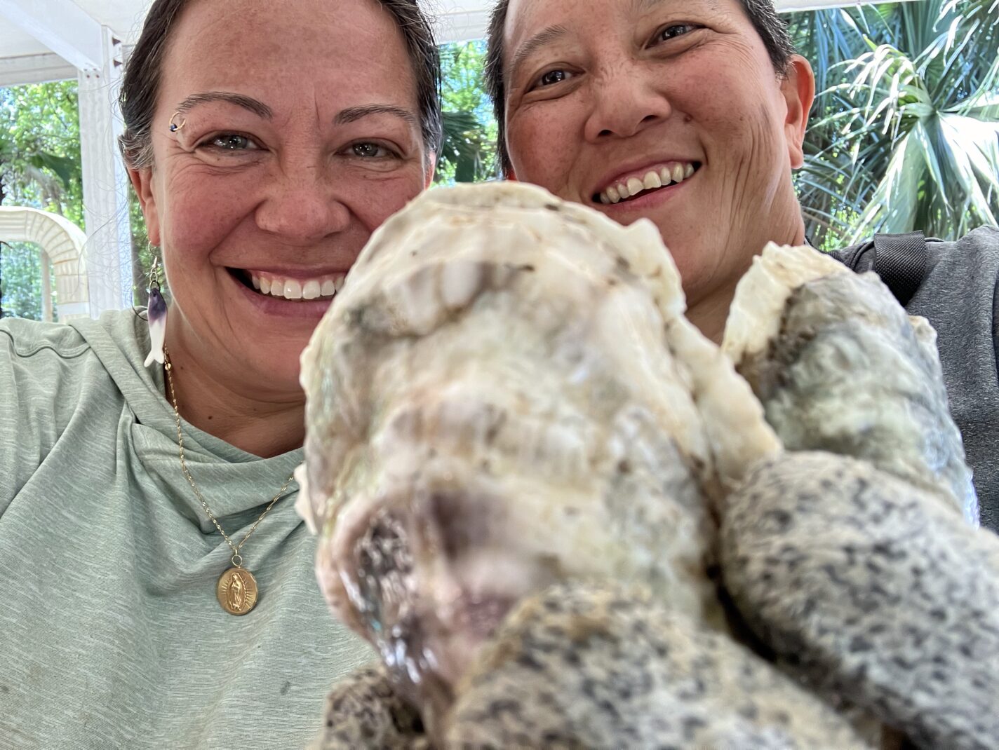 Two oyster farmers from Water if Life Oysters are wearing gray sweat shirts are smiling and standing together. One has a glove on and is holding a few oyster very close in the foreground. Sun and palm trees shine behind them.