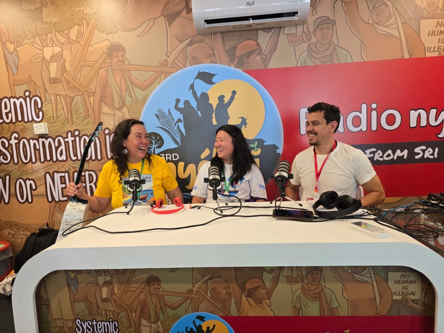 Melanie Brown, a NAMA board member, salmon fisher, and musician, along with NAMA communications director Feini Yin, are interviewed at a white table during the Nyéléni Forum in Sri Lanka. A massive, colorful painting of farmers and others is shown on the wall behind them.