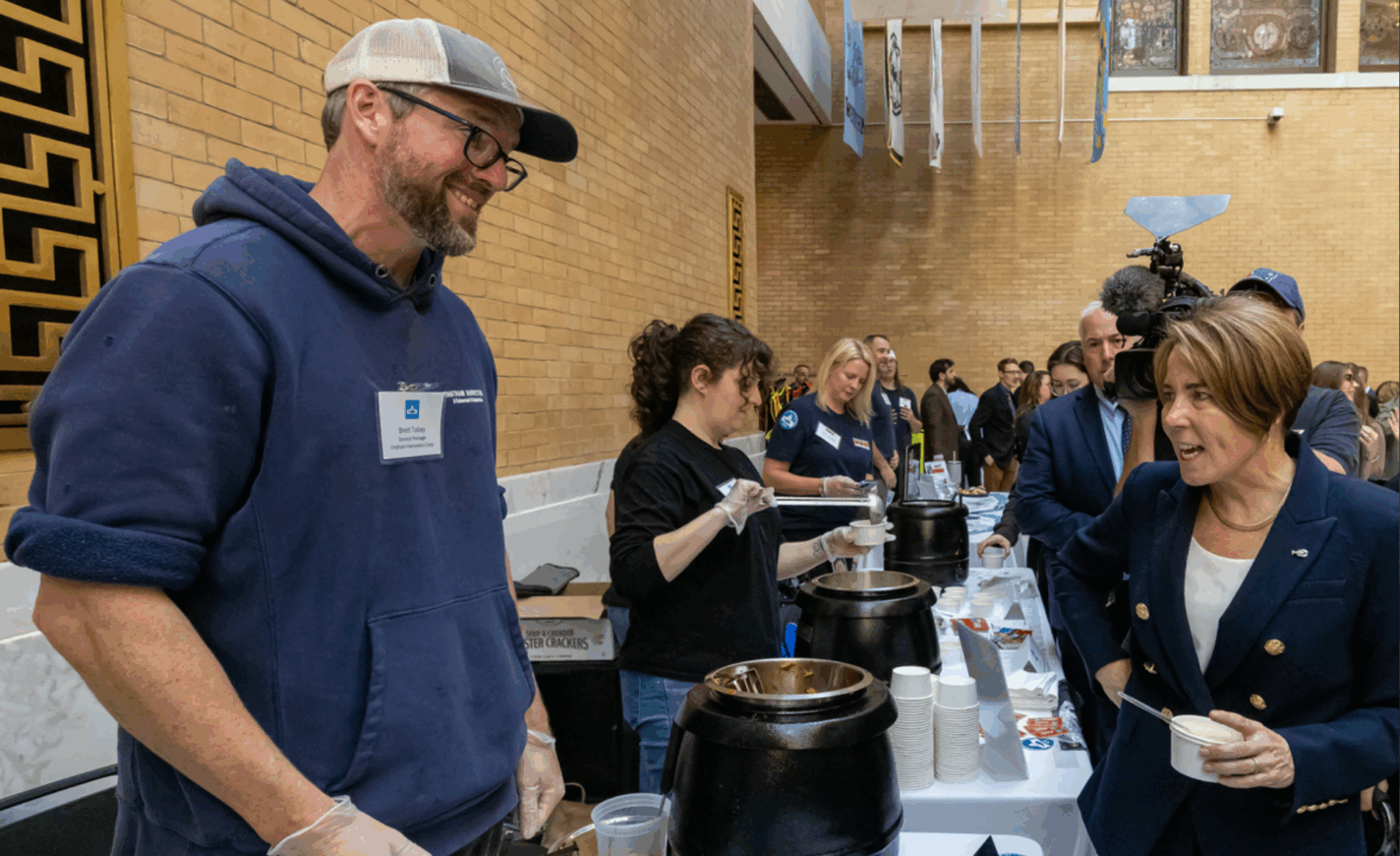 A very tall Brett Tolley wears a navy blue sweatshirt, a baseball cap, and serving gloves as he chats with Massachusetts Governor Maura Healey, whom Brett served Chatham Harvester's BBQ-style skate fish.