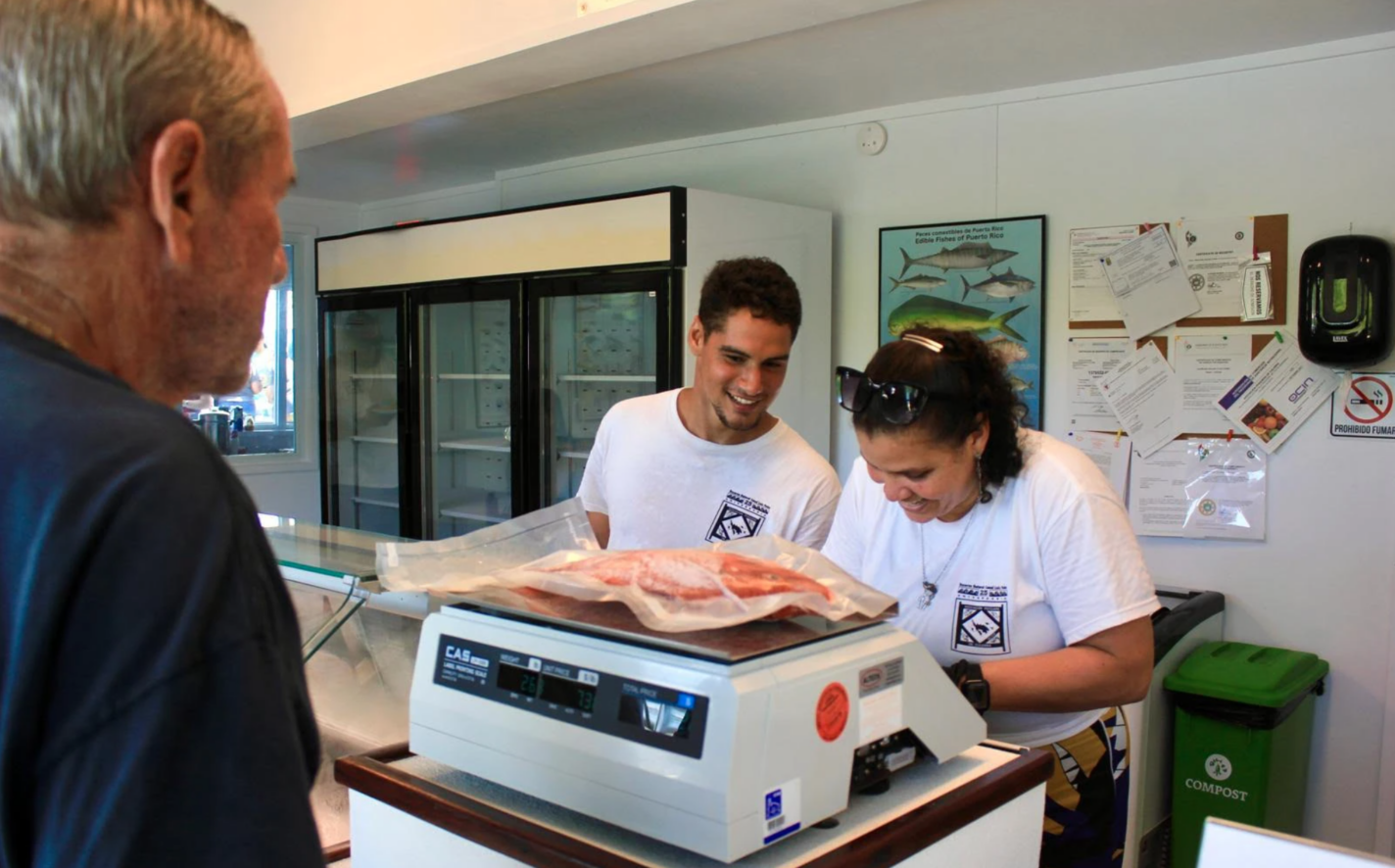 Two workers wearing white t-shirts stand behind a counter weighing fish and smiling as an elderly person on the other side of the counter orders fish.