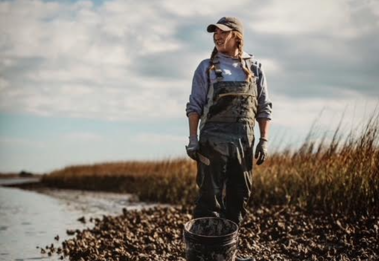 Fisherwoman Ana Shellem stands in a marshy area with a partly cloudy sky behind her. She is wearing a baseball cap and suspenders and is smiling. A muddy bucket sits in front of her.