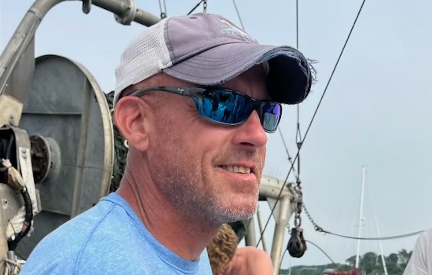Doug Feeney stands on a dock. He wears sunglasses and a gray baseball cap.