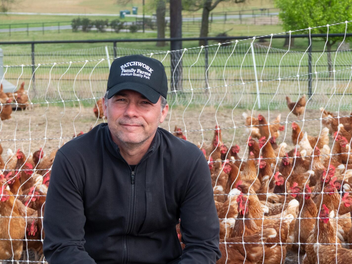 NFFC director Tim Gibbons wears a black shirt and hat, sits in front of several chickens that are behind a fence.